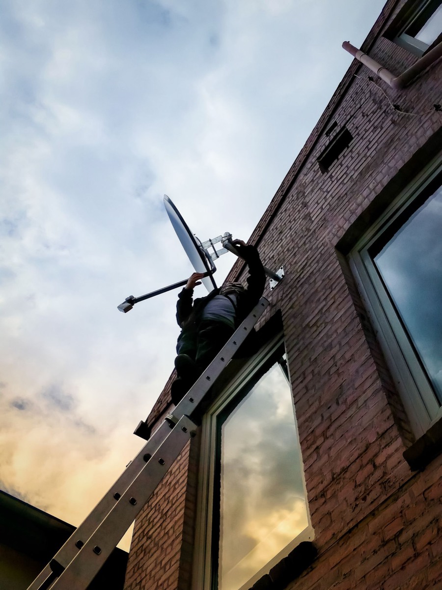 Technician installing a satellite dish high on a wall at dusk